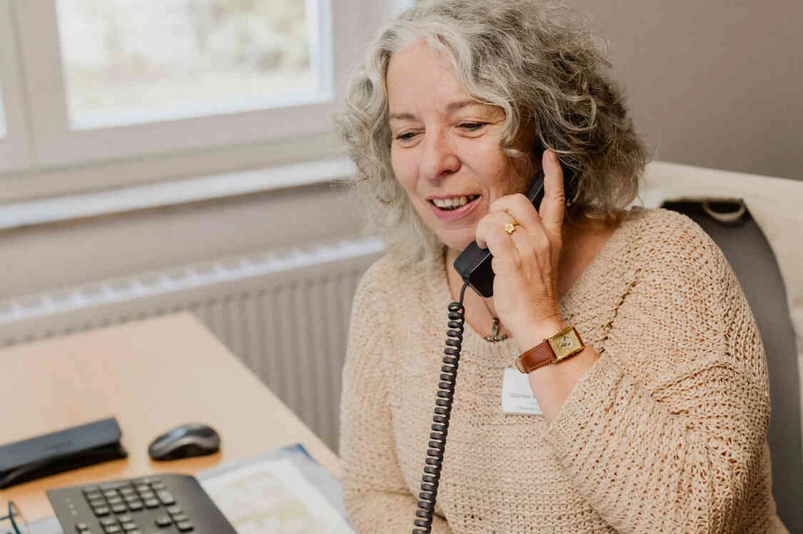 Mitarbeiterin mit Namensschild telefoniert am Schreibtisch neben Tastatur und Unterlagen