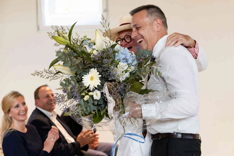 Zwei M&auml;nner umarmen sich bei der Er&ouml;ffnung, einer h&auml;lt einen Blumenstrau&szlig;.