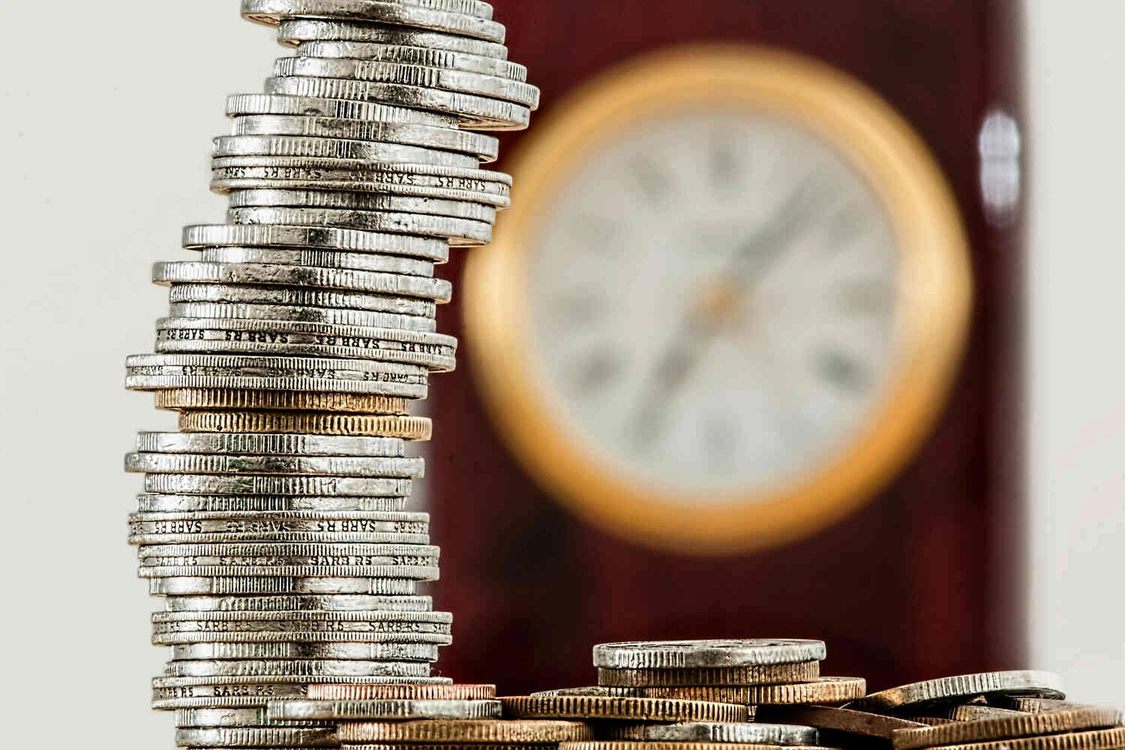 M&uuml;nzstapel vor einer unscharfen Uhr als Symbol f&uuml;r Zeit und Geld