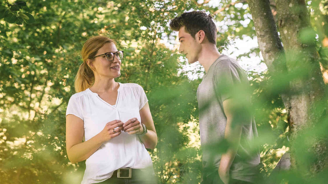 Frau mit Brille und junger Mann stehen im Gespr&auml;ch zwischen B&auml;umen