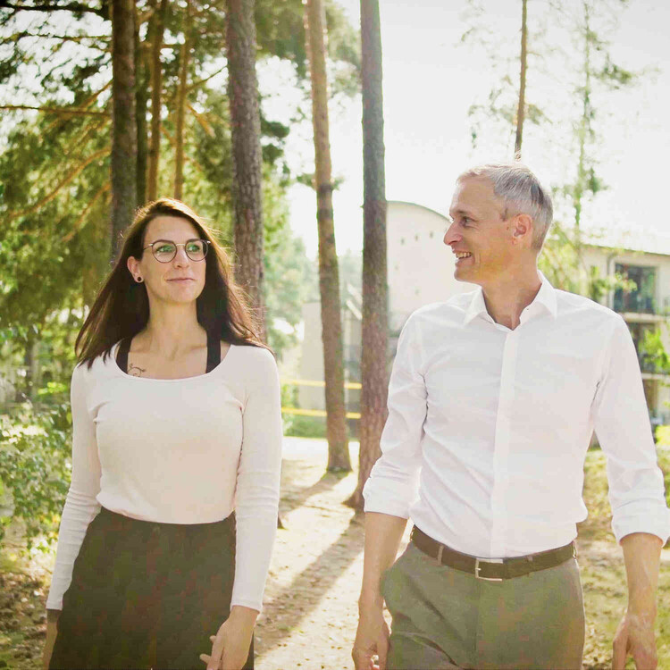 Frau und Mann gehen durch eine gr&uuml;ne Au&szlig;enanlage vor einem Klinikgeb&auml;ude