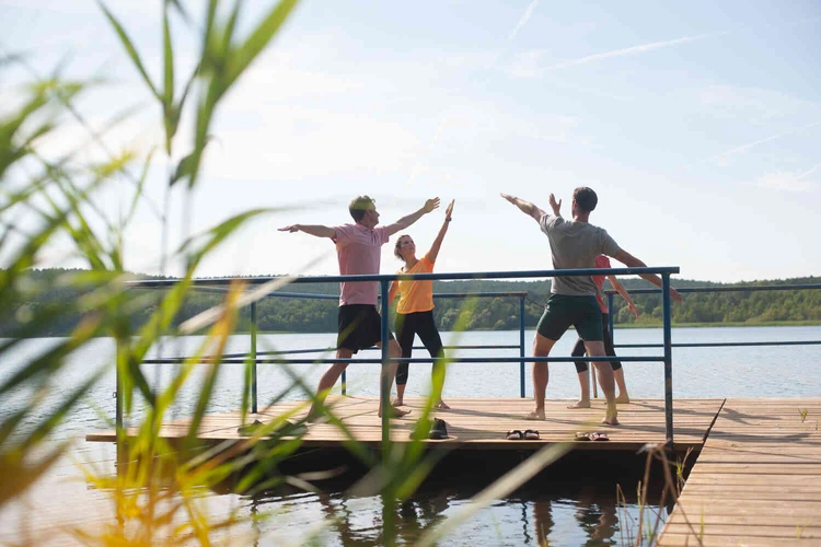 Drei Personen machen Bewegungs&uuml;bungen auf einem Steg am See