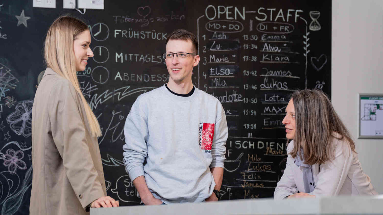 Zwei junge Erwachsene sprechen mit einer sitzenden Frau vor einer Tafel mit Tagesplan