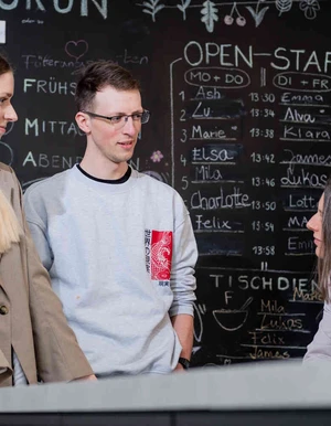 Zwei junge Erwachsene sprechen mit einer sitzenden Frau vor einer Tafel mit Tagesplan