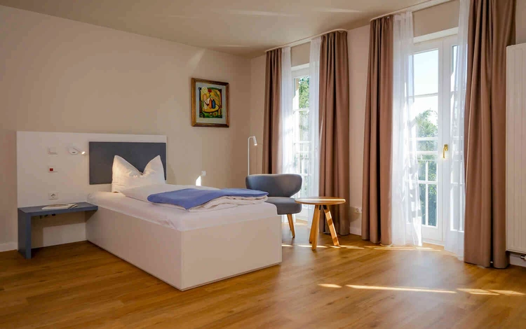 Helles Patientenzimmer mit Bett, Sessel und Balkonfenstern