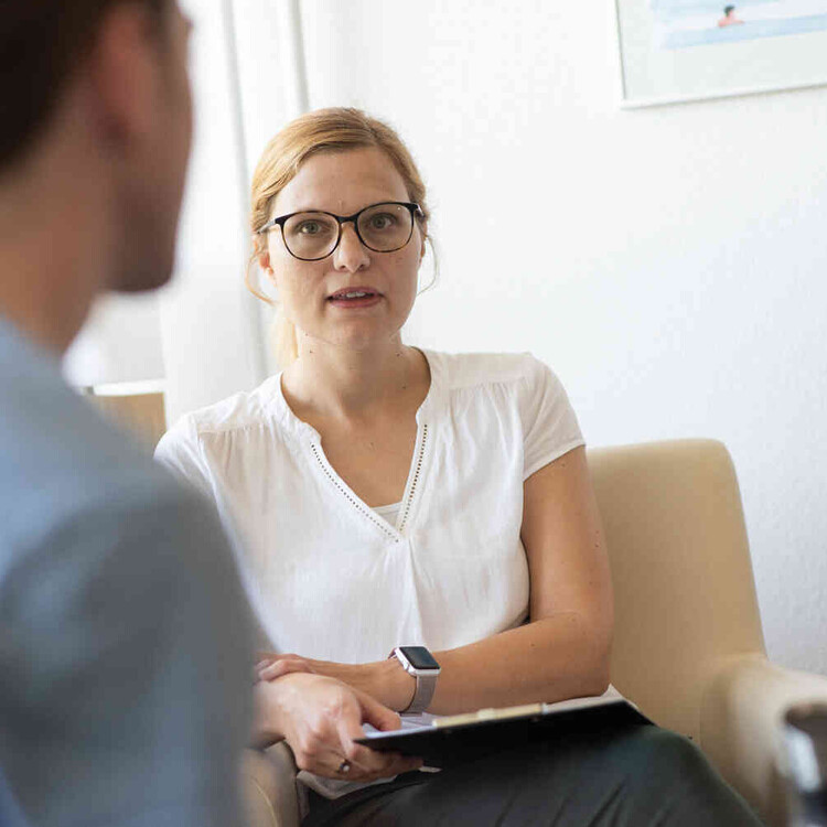 Frau mit Brille sitzt in einem Gespr&auml;ch und h&auml;lt ein Klemmbrett