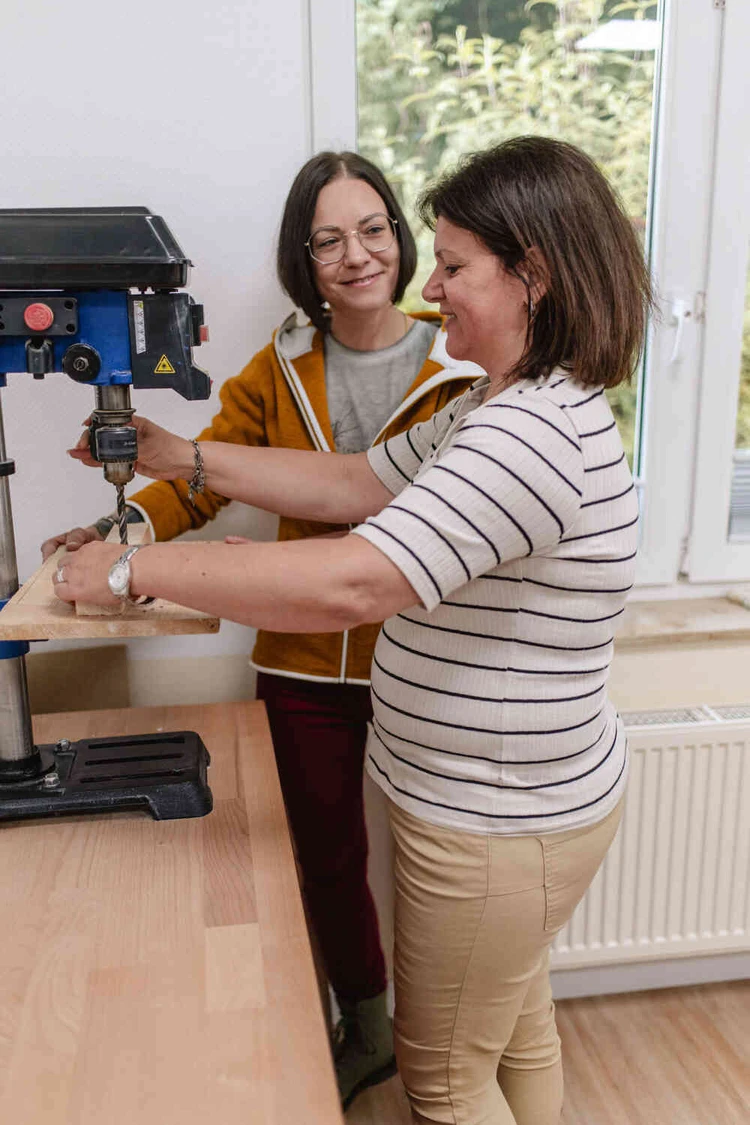 Zwei Frauen l&auml;cheln beim Bohren eines Holzst&uuml;cks an einer Standbohrmaschine