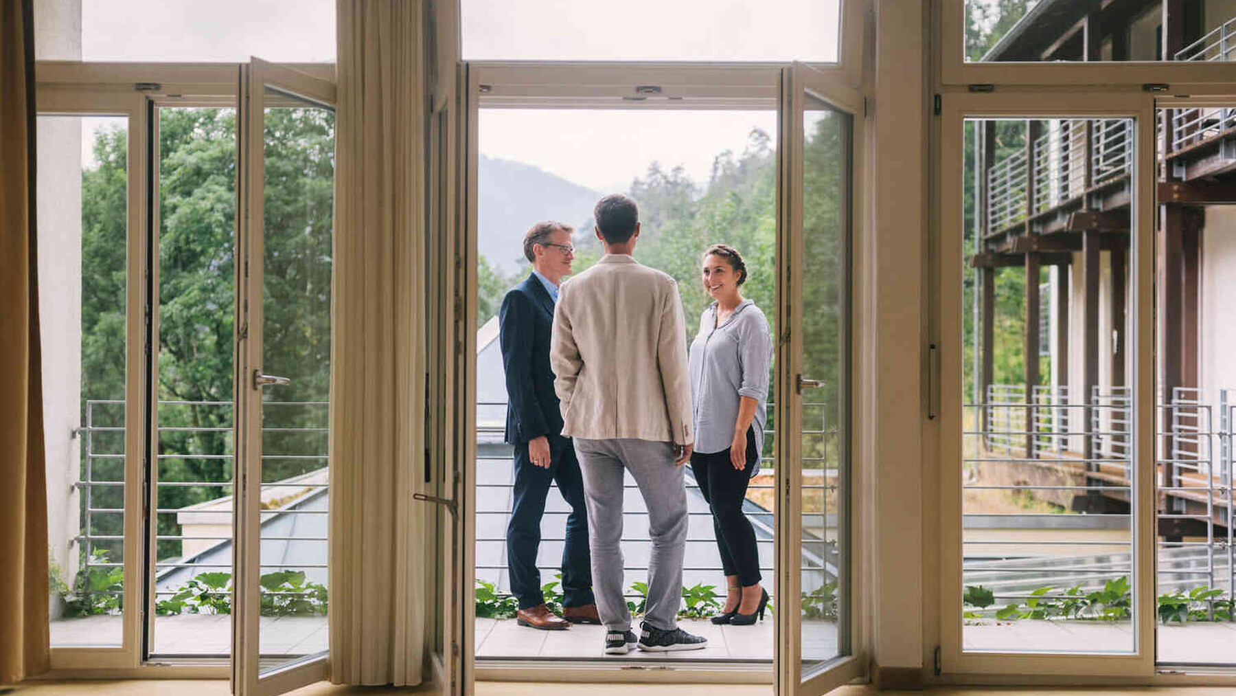 Drei Personen stehen im Gespr&auml;ch auf einem Balkon mit Blick ins Gr&uuml;ne