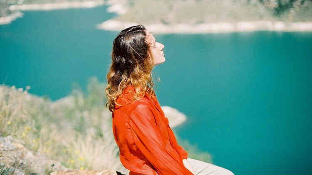 Frau in roter Bluse sitzt am See und blickt in die Sonne