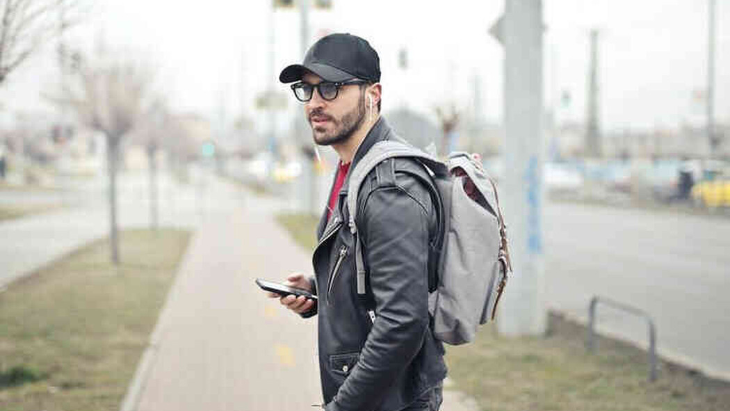 Mann mit Kappe, Rucksack und Smartphone steht auf einem Gehweg