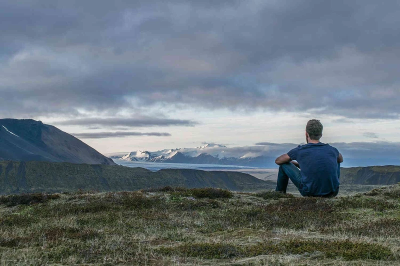 Mann sitzt allein vor einer weiten Berglandschaft unter bew&ouml;lktem Himmel