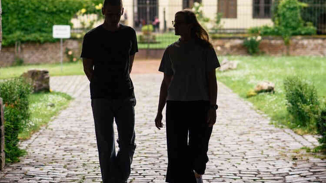 Zwei Personen gehen im Schatten &uuml;ber einen gepflasterten Weg vor einem Geb&auml;ude